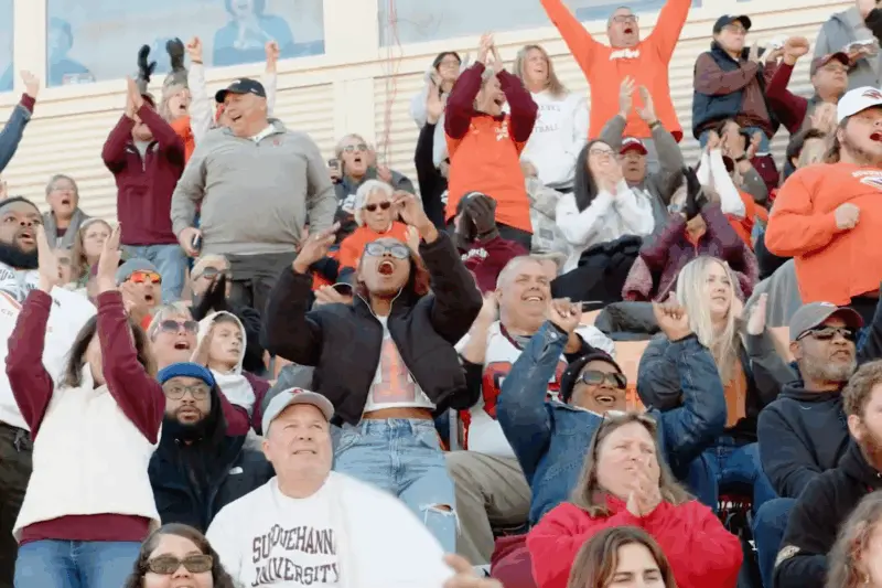 A diverse crowd at a sports event, wearing various team apparel, cheering enthusiastically with raised arms. The scene captures a mix of emotions, including joy and excitement, as spectators engage with the game.