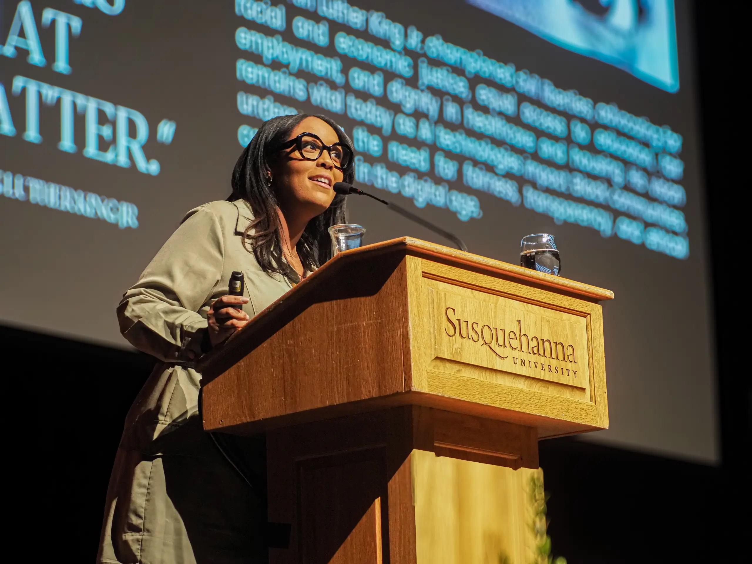 A woman speaking at a podium with "Susquehanna University" on the front. She is wearing glasses and holding a microphone. Behind her is a large screen with text, likely part of a presentation or lecture.