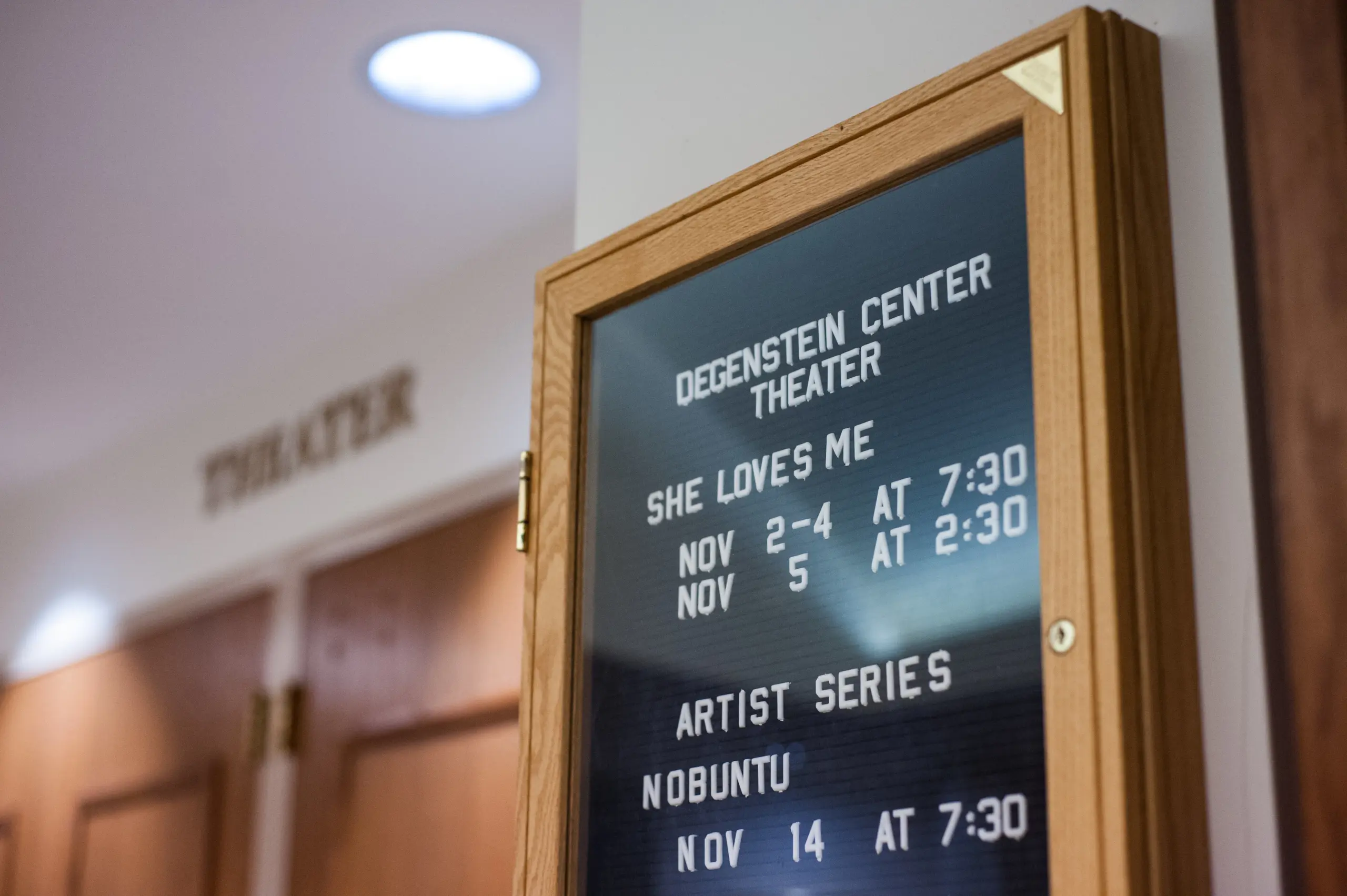 A theater sign lists upcoming events: "She Loves Me" on Nov 2-4 at 7:30 PM and Nov 5 at 2:30 PM, followed by "Artist Series Nobuntu" on Nov 14 at 7:30 PM. The sign is in a wooden frame, mounted on a wall near closed doors.