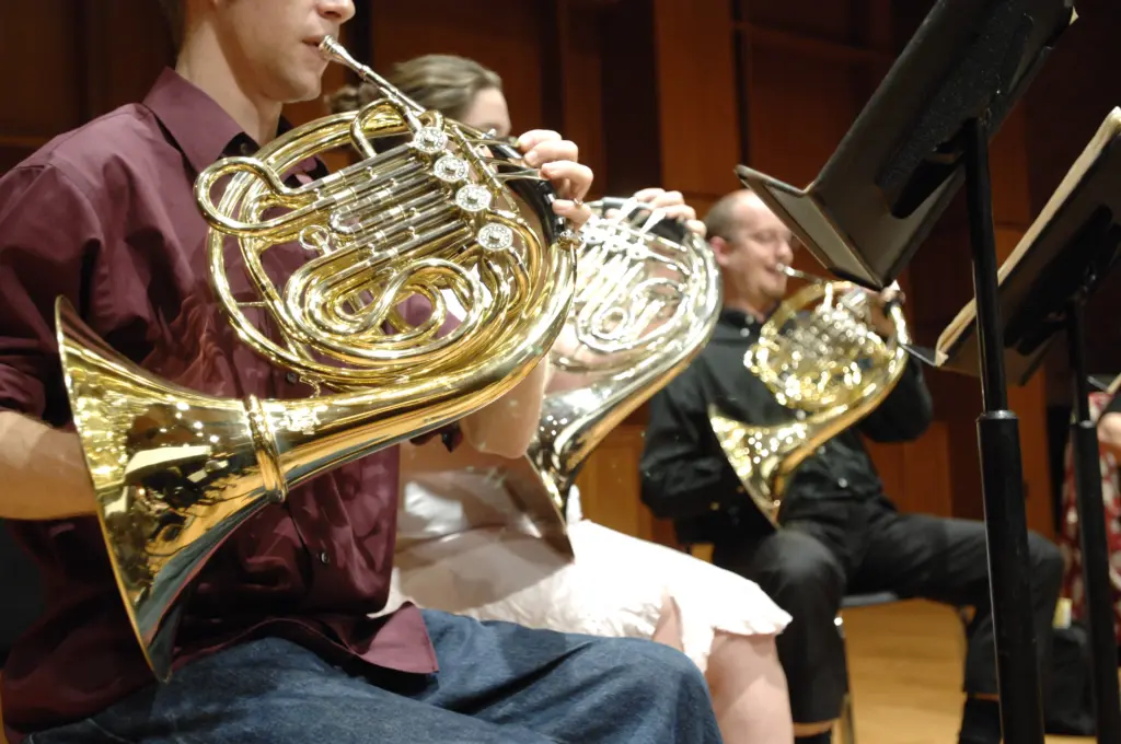 Three musicians skillfully play French horns in an orchestral setting, possibly gearing up for music festivals. They are seated in a row with sheet music on stands, poised against a wood-paneled backdrop that evokes the ambiance of a concert hall or rehearsal space.