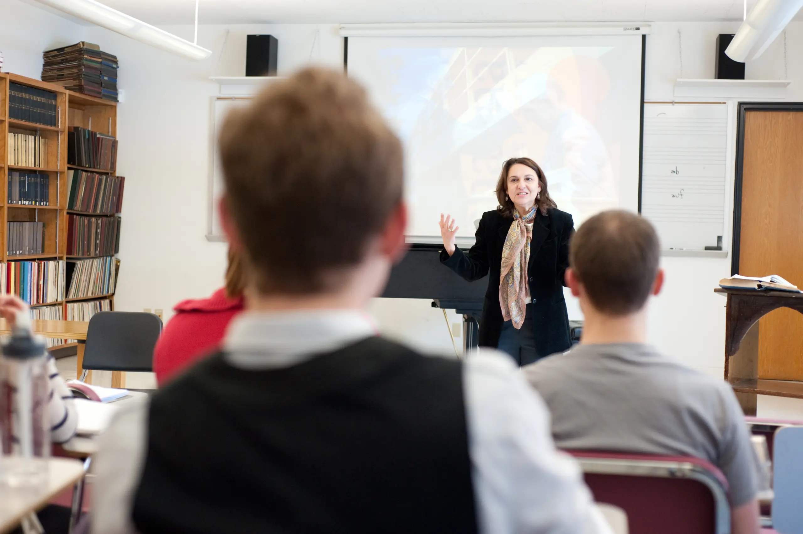 A woman is giving a lecture in a classroom, gesturing with her hands. She stands in front of a projector screen with a blurred image. Several students are sitting and facing her, listening attentively. Bookshelves and a whiteboard are in the background.