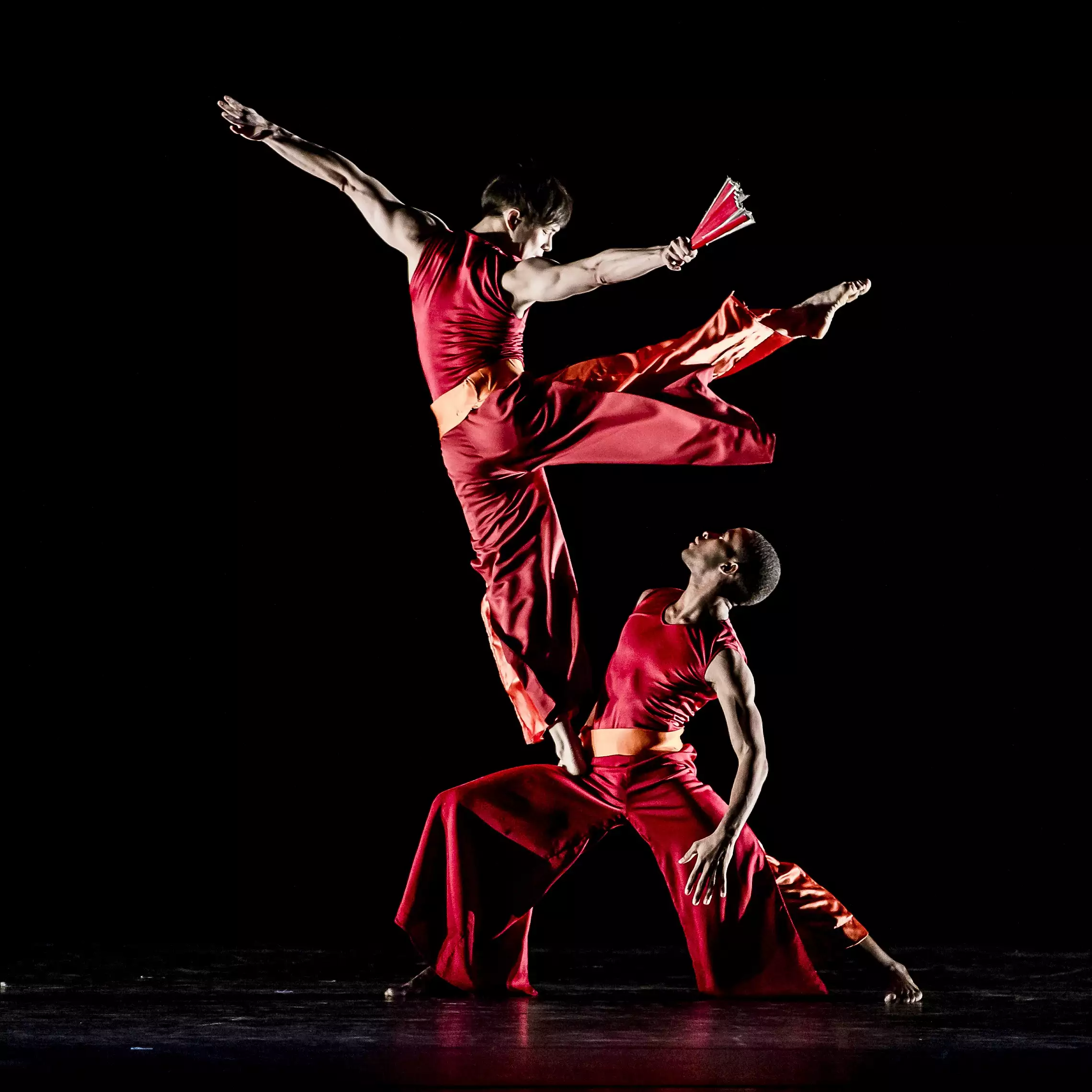 Two dancers in red costumes perform an elegant aerial move against a black background. One dancer leaps with a fan in hand, while the other supports them, showcasing their strength and balance.