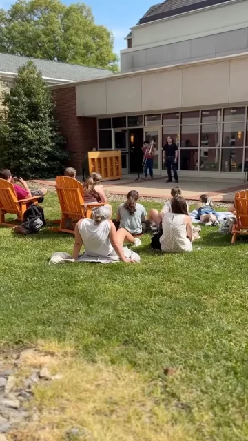 A group of people sits on a grassy lawn and wooden chairs, enjoying an outdoor piano performance near the Student Health & Wellness center. Another person stands nearby. The scene is bright and sunny, with trees and windows framing the background.