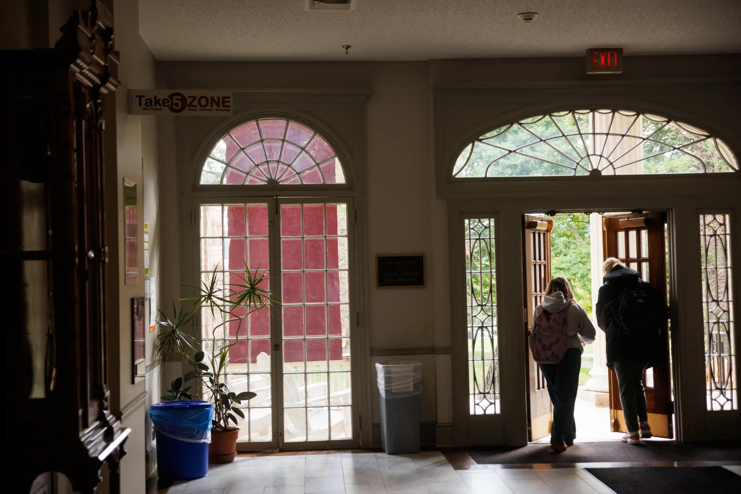 Two people with backpacks exit through an open double door of an Upper-Level Housing building. Indoor lighting reveals a plant, a blue trash bin, and a "Take 5 Zone" sign. Arched windows and outdoor greenery are visible in the background.