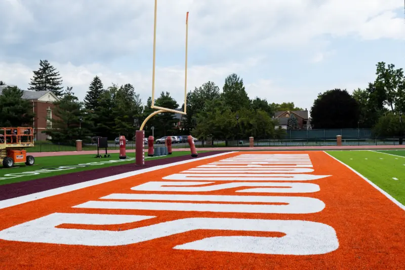 Close-up of a football field's end zone, featuring bright orange turf with the word "Indians" in large white letters, embodies the spirit of athletics and recreation. Yellow goalposts stand against a partly cloudy sky, framed by trees along the perimeter.