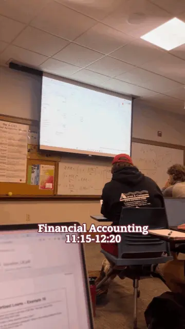 In a classroom at the Sigmund Weis School of Business, students are seated facing a projector displaying a spreadsheet. The chalkboard behind them features various formulas, and text reads "Financial Accounting 11:15-12:20." One student stands out in a red cap and sweatshirt.