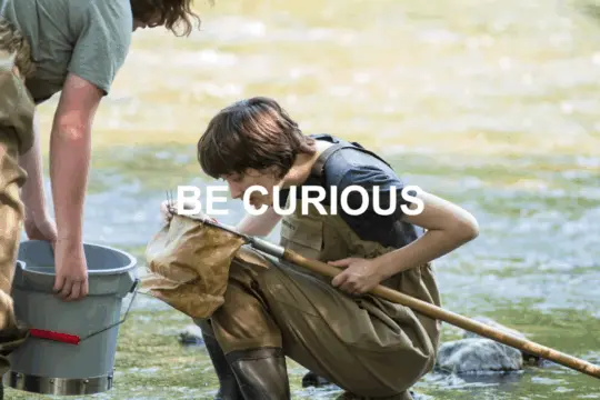 Two people in waders examine a net by the sunlit river, one holding a small gray bucket. The scene captures the spirit of Pre-College Summer Programs that inspire students to be curious and explore nature up close. Text overlay reads, "BE CURIOUS.