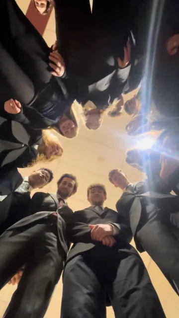 A group of people in formal attire, presumably from the Sigmund Weis School of Business, stand in a circle, looking down at the camera. The ceiling and a light fixture are visible above them.