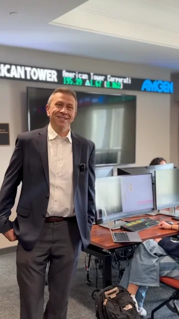 A person in a suit, smiling confidently beneath a digital stock ticker, exemplifies the professional excellence fostered by the Sigmund Weis School of Business. In the background, computer stations with engaged individuals highlight a commitment to cutting-edge analytics and real-world applications.