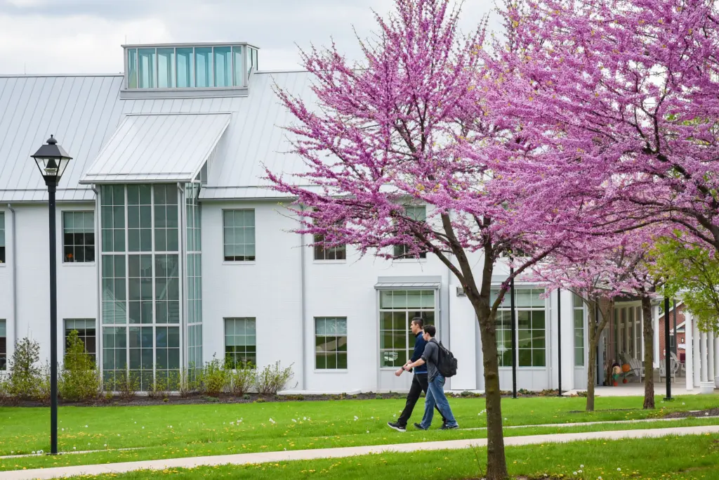 Two people walk on a path lined with blossoming pink trees, while the modern white building with large windows showcases its commitment to sustainability initiatives. The vibrant green grass adds a touch of nature's elegance beneath a cloudy sky.