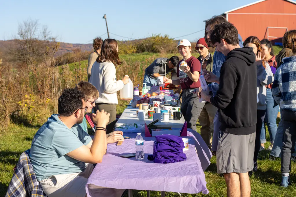 A group of people gathers outdoors on a sunny day around tables covered with purple cloths and various items. Some are seated, while others stand, chatting about local sustainability initiatives. Trees and a red building create a charming backdrop for the lively discussion.