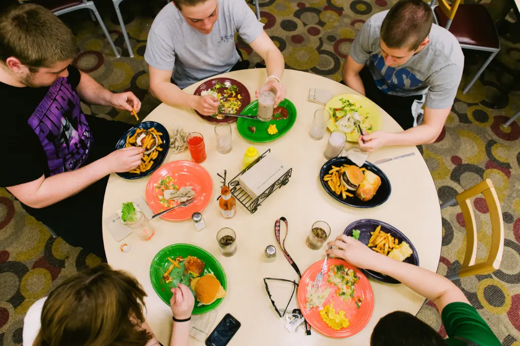 Five people sitting around a round table, enjoying a meal together. Plates with sustainably-sourced burgers, fries, salads, and various drinks are spread out. The floor boasts a patterned design.