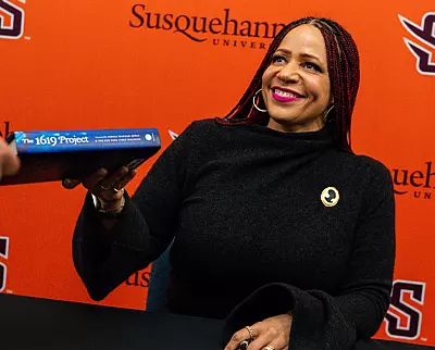 A smiling woman with long braids sits at a table, handing a copy of "The 1619 Project" to someone. The background features the Susquehanna University logo and name on an orange wall.