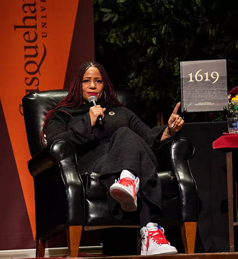 A woman with long braids sits in a black armchair on stage, speaking and gesturing beside a book titled "1619." She wears a dark outfit and white sneakers with red accents. A Susquehanna University banner is in the background.