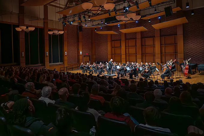 A symphony orchestra performs on stage in a concert hall, with a conductor leading the musicians. The audience is seated in rows, attentively watching the performance under warm stage lighting.