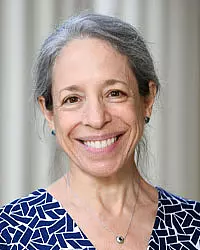 Smiling woman with long gray hair, wearing a blue and white patterned top and a small pendant necklace, standing in front of a softly blurred background.