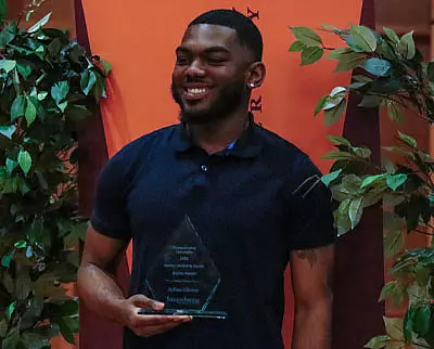 A smiling man in a dark shirt holds a clear, triangular award in front of an orange backdrop with green leafy plants on either side.