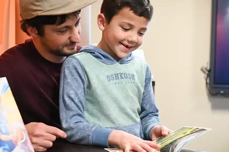 An adult and a smiling young boy sit together at a table, reading a book. Another illustrated book stands upright nearby. The adult looks at the boy, while the boy points at the open book’s pages.