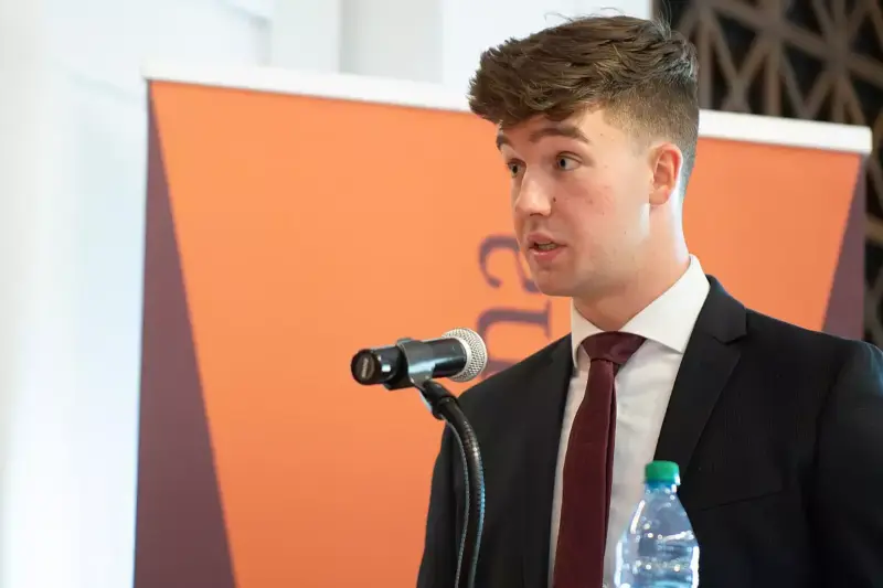 A young man in a suit and tie speaks into a microphone at a podium, with a bottle of water in front of him and an orange banner in the background.