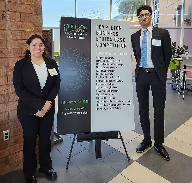 Two professionally dressed students stand beside a large sign for the "Templeton Business Ethics Case Competition" at Stetson University. The sign lists event dates, sponsors, and participating institutions.