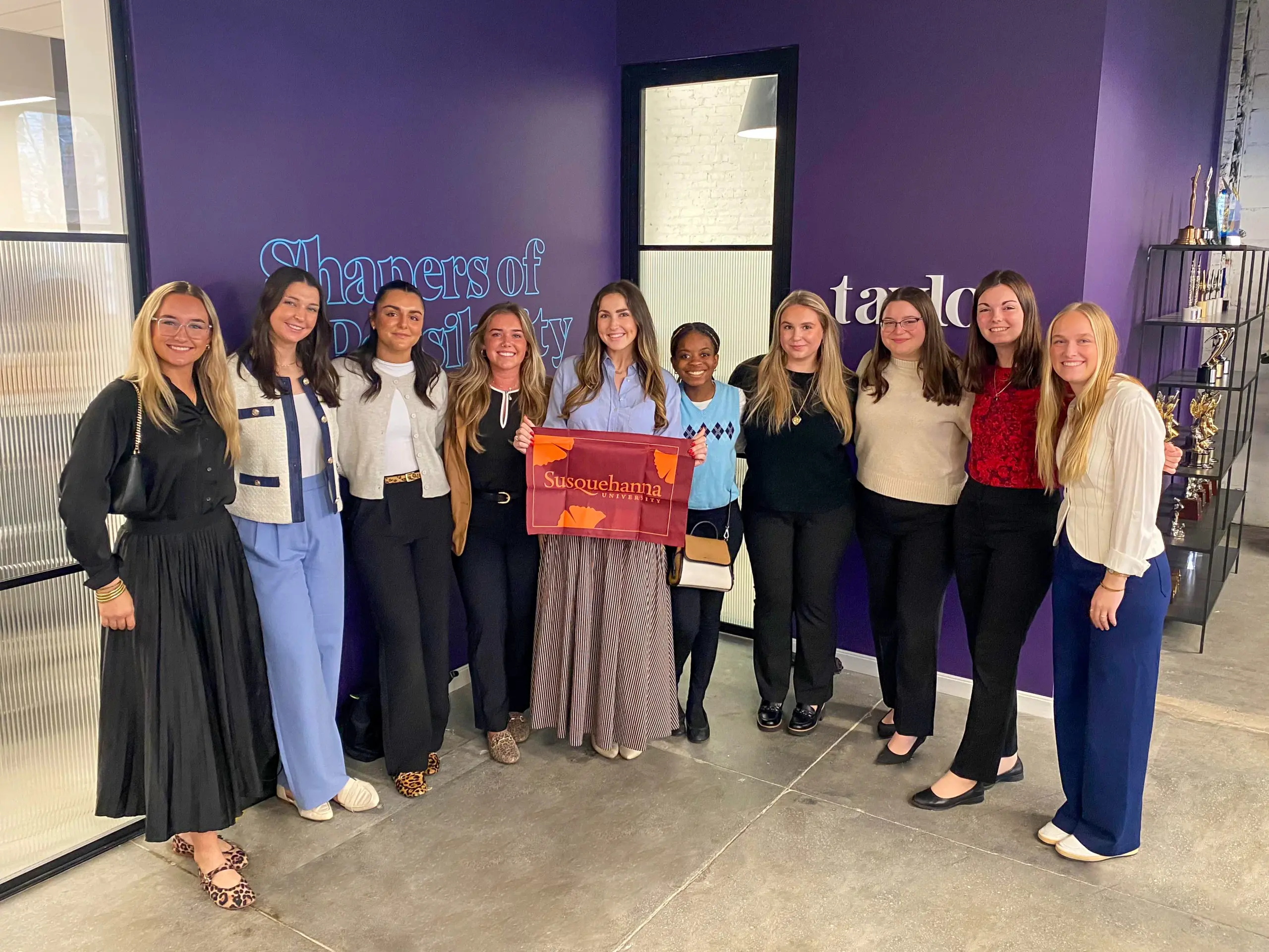 Ten women stand together indoors in front of a purple wall, smiling at the camera. One woman in the center holds a red and orange “Susquehanna” pennant, highlighting Student & Alumni Networking in a business casual setting.