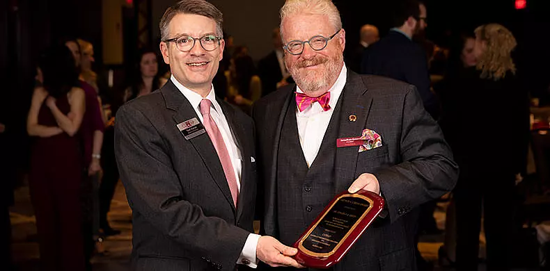 Our Leadership shines as two men in suits and glasses smile while holding an award plaque together at an indoor event, with other people mingling in the blurred background.