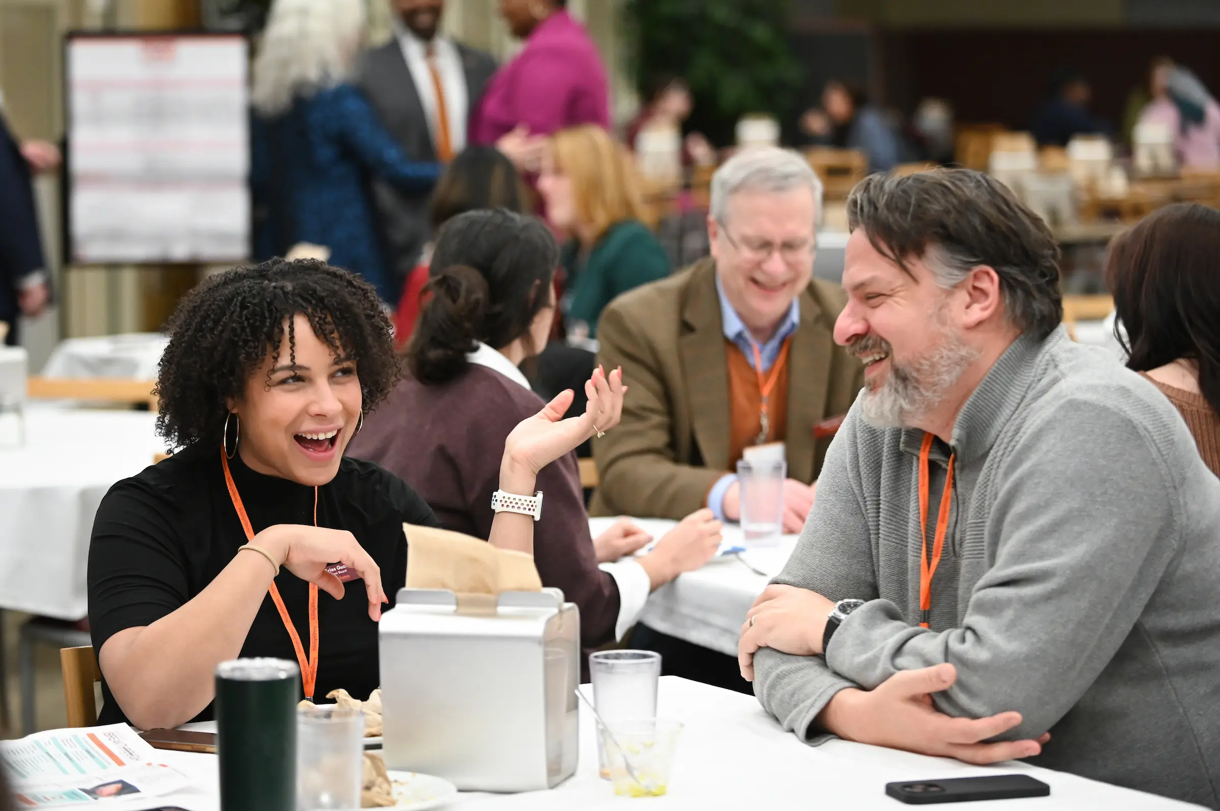 Two people sit at a table, laughing and talking with each other at what appears to be a conference or Giving Societies event. Other attendees are conversing in the background. Both are wearing orange lanyards.