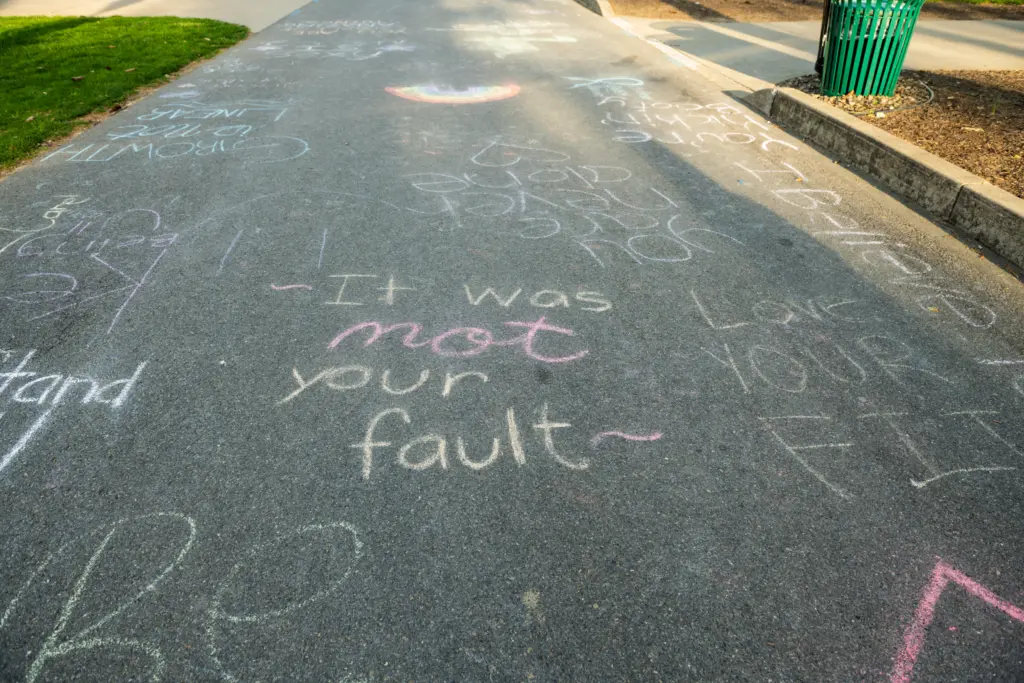 A paved path covered in colorful chalk messages, including a large, prominent phrase in the center that reads, "It was not your fault," along with other supportive and positive words and drawings promoting safety on campus.
