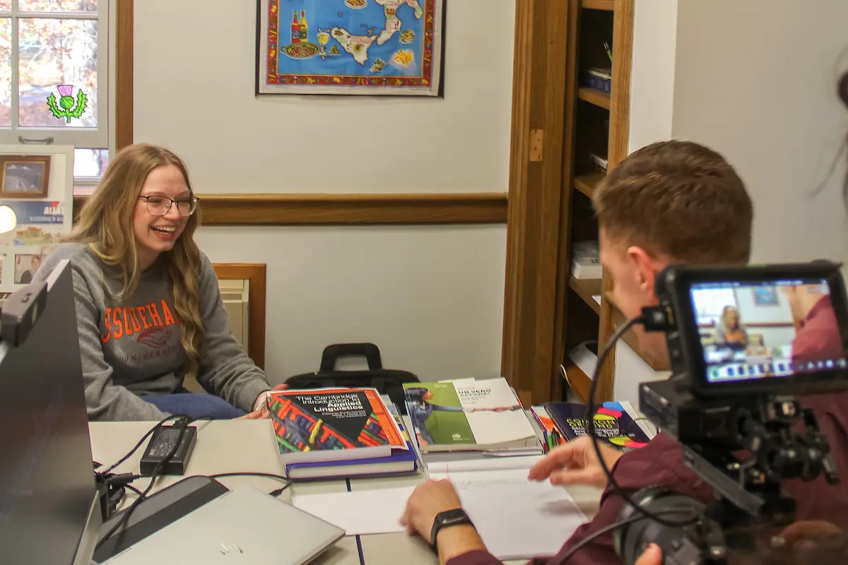 A young woman with glasses sits at a desk, smiling, while a man interviews her on camera. Surrounded by books and a laptop, she discusses academics in a small office with a map on the wall.