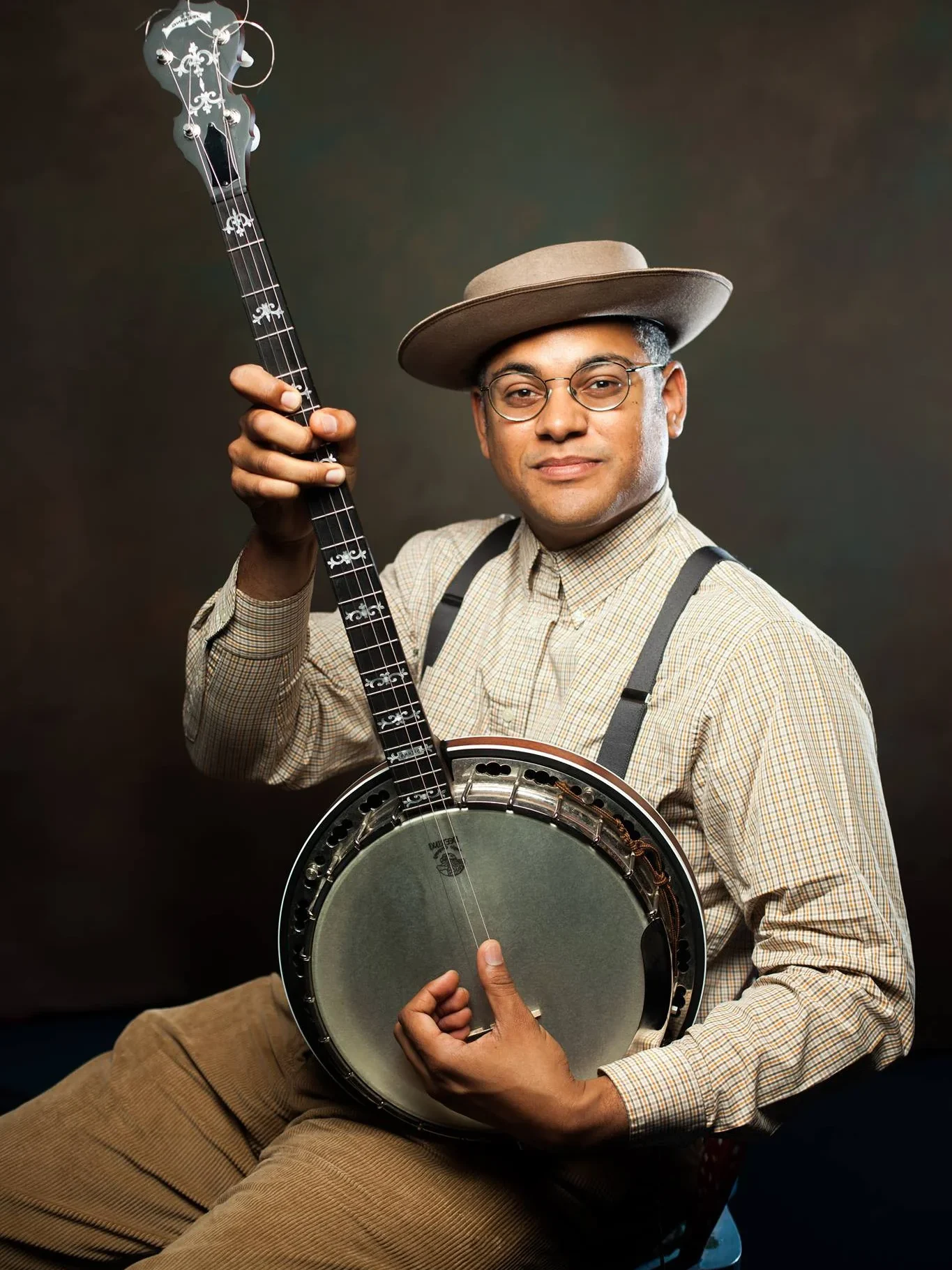 A person wearing glasses, a brown hat, suspenders, and a plaid shirt sits on a stool, holding up a banjo and looking at the camera against a dark background, celebrating the spirit of Community Access through music.