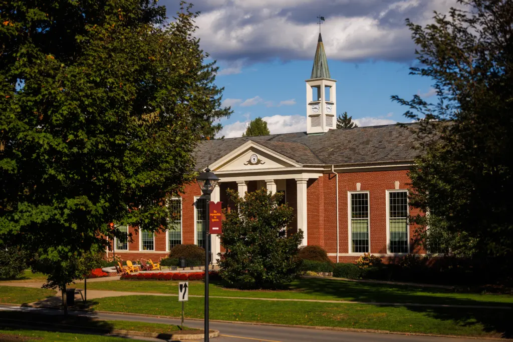 A red brick academic building with tall white columns and a cupola stands behind trees on a sunny day, with a manicured lawn and a lamppost in front.