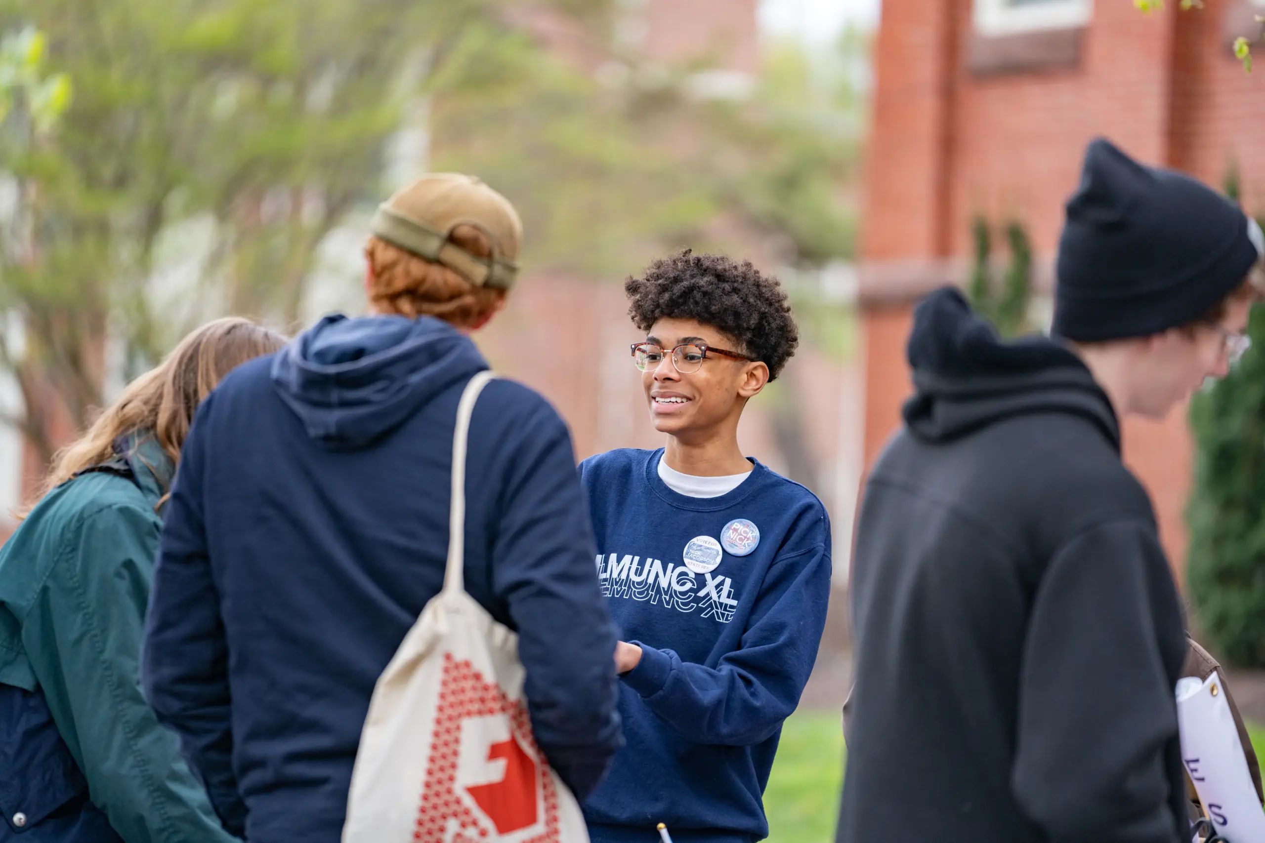 A young person wearing glasses and a navy sweater smiles while talking to others outdoors. Three people face away, and the group stands near leafy trees and a red brick building.