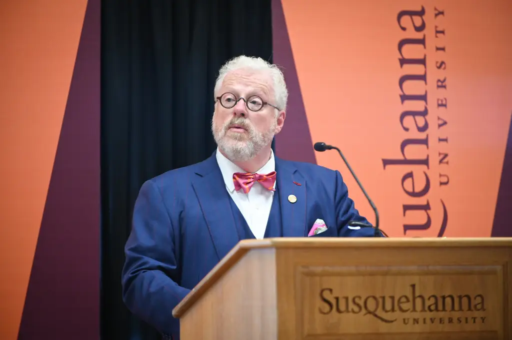 A man with white hair and a beard, representing Our Leadership, wears glasses, a blue suit, and a red bow tie as he speaks at a podium labeled "Susquehanna University" with a university banner in the background.