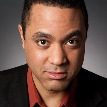 A man with short curly hair and medium skin tone, wearing a dark blazer over a red collared shirt, looks directly at the camera with a neutral expression. The softly lit, dark background hints at themes of community access and inclusion.