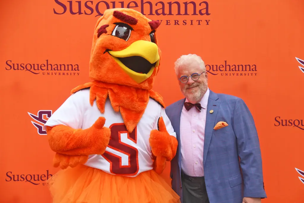 Our Leadership shines as a person in a blue suit and bow tie stands smiling next to an orange bird mascot in front of a Susquehanna University backdrop. The mascot gives a thumbs up, wearing an "S" shirt and orange tutu.