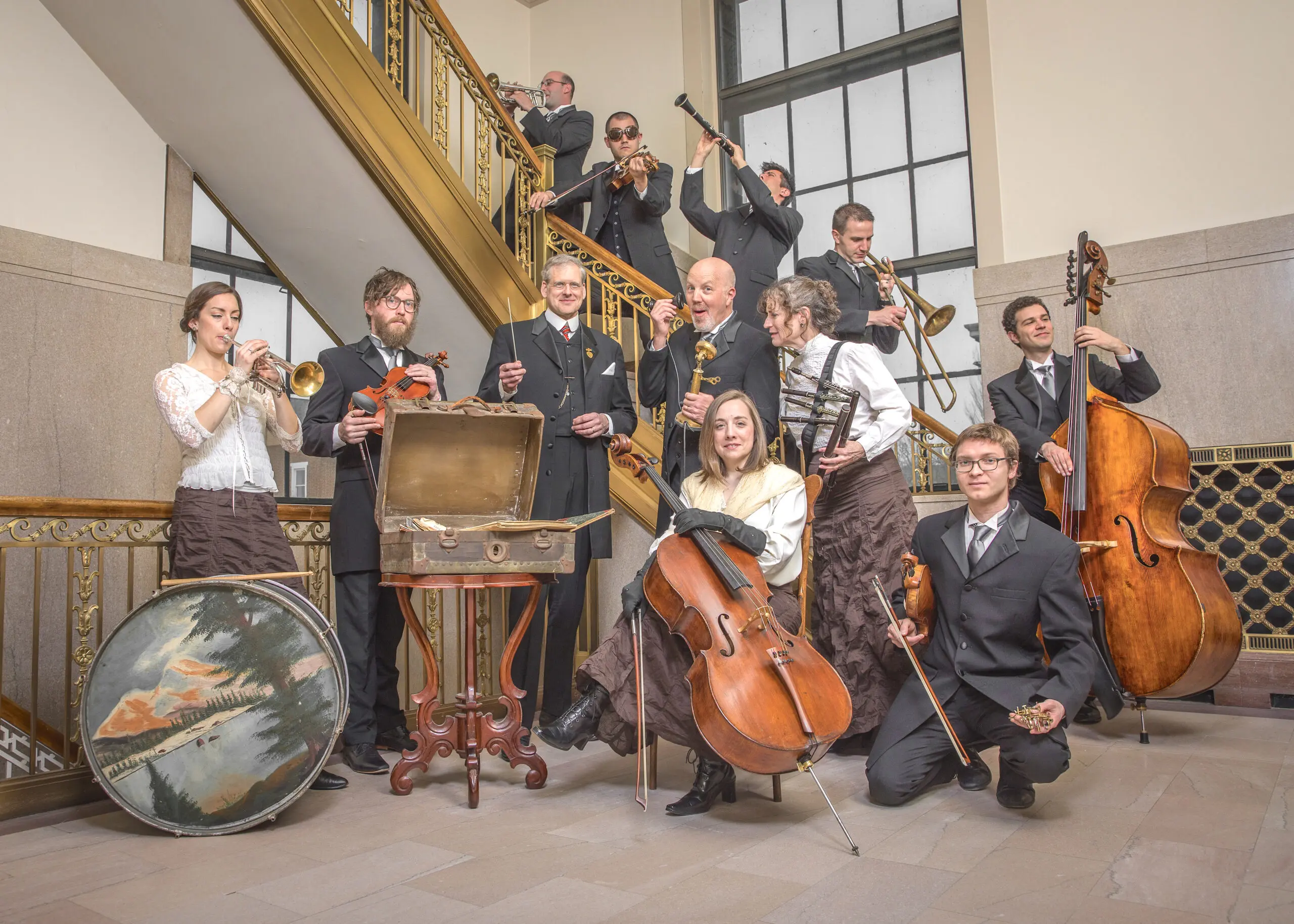 A group of eleven musicians in formal attire pose by a staircase, holding instruments like cello, violin, trumpet, flute, clarinet, and drums, highlighting Community Access to music with an open chest in front of them.