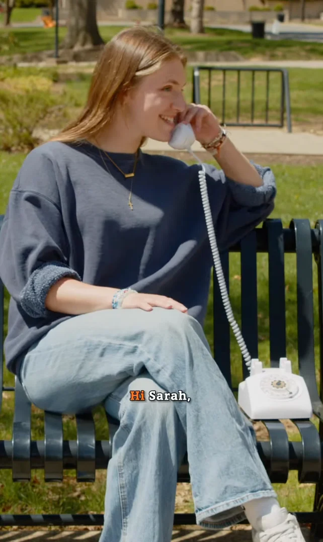 A young woman sits on a park bench, smiling while talking on a white rotary phone. She wears a loose blue sweater and light jeans, celebrating her commencement moment. The subtitle says, "Hi Sarah," with "Hi" highlighted in orange.