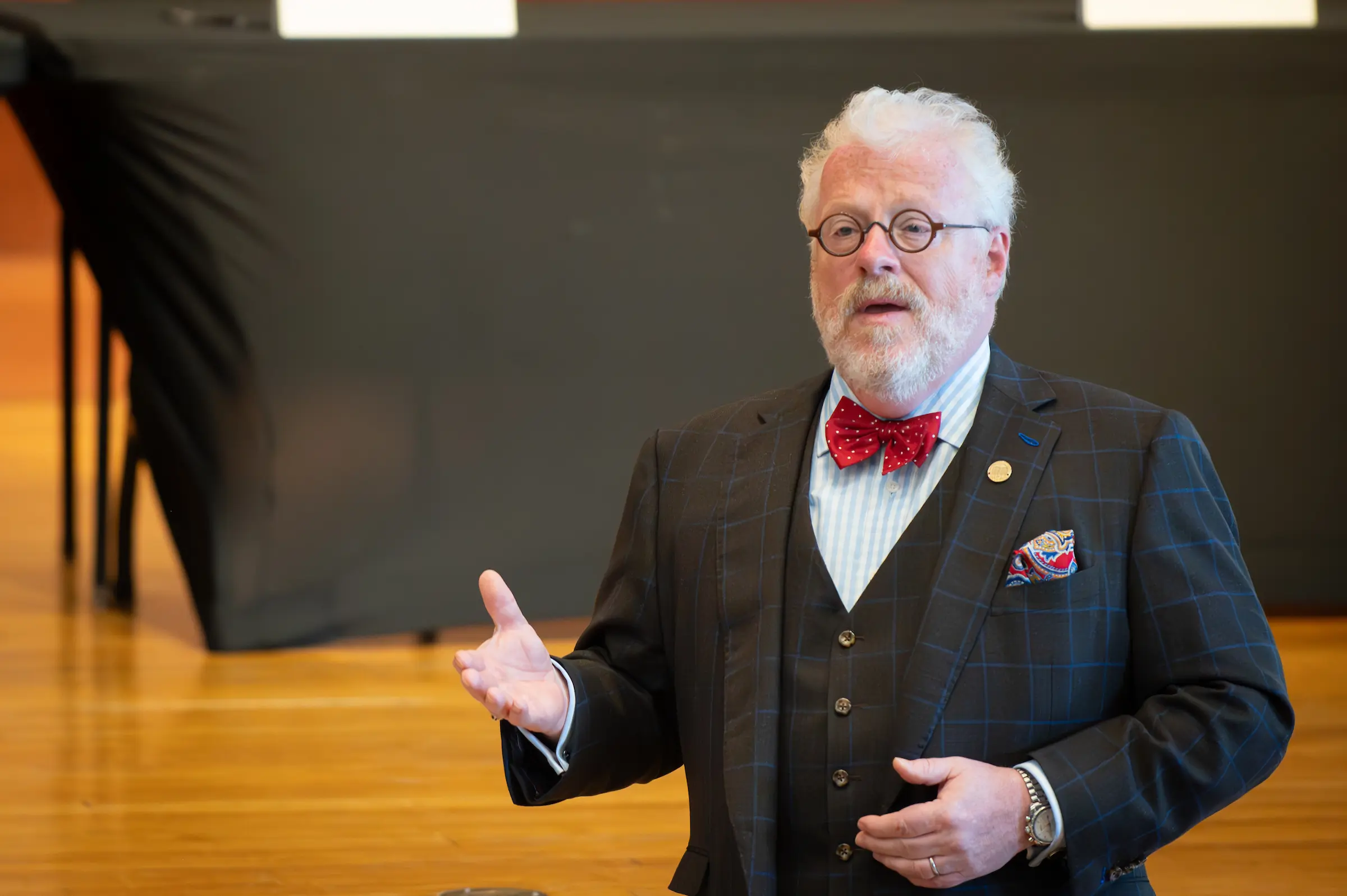 An older man with white hair and a beard, wearing round glasses, a dark plaid suit, red bow tie, and patterned pocket square, gestures with one hand while speaking indoors—an inspiring figure among Our Leadership.