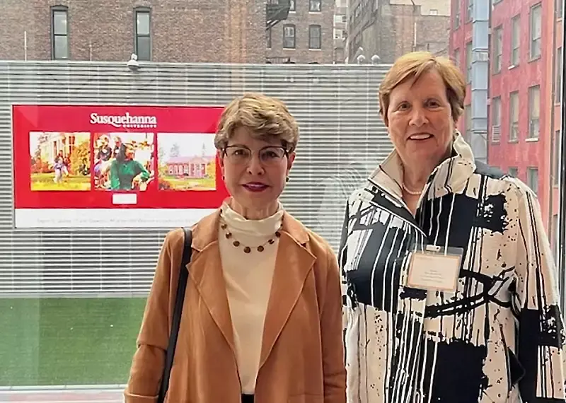 Two women stand indoors by a large window with city buildings in the background, connecting during a Student & Alumni Networking event. One wears a tan jacket and glasses; the other sports a black and white patterned jacket. A Susquehanna University sign is visible behind them.