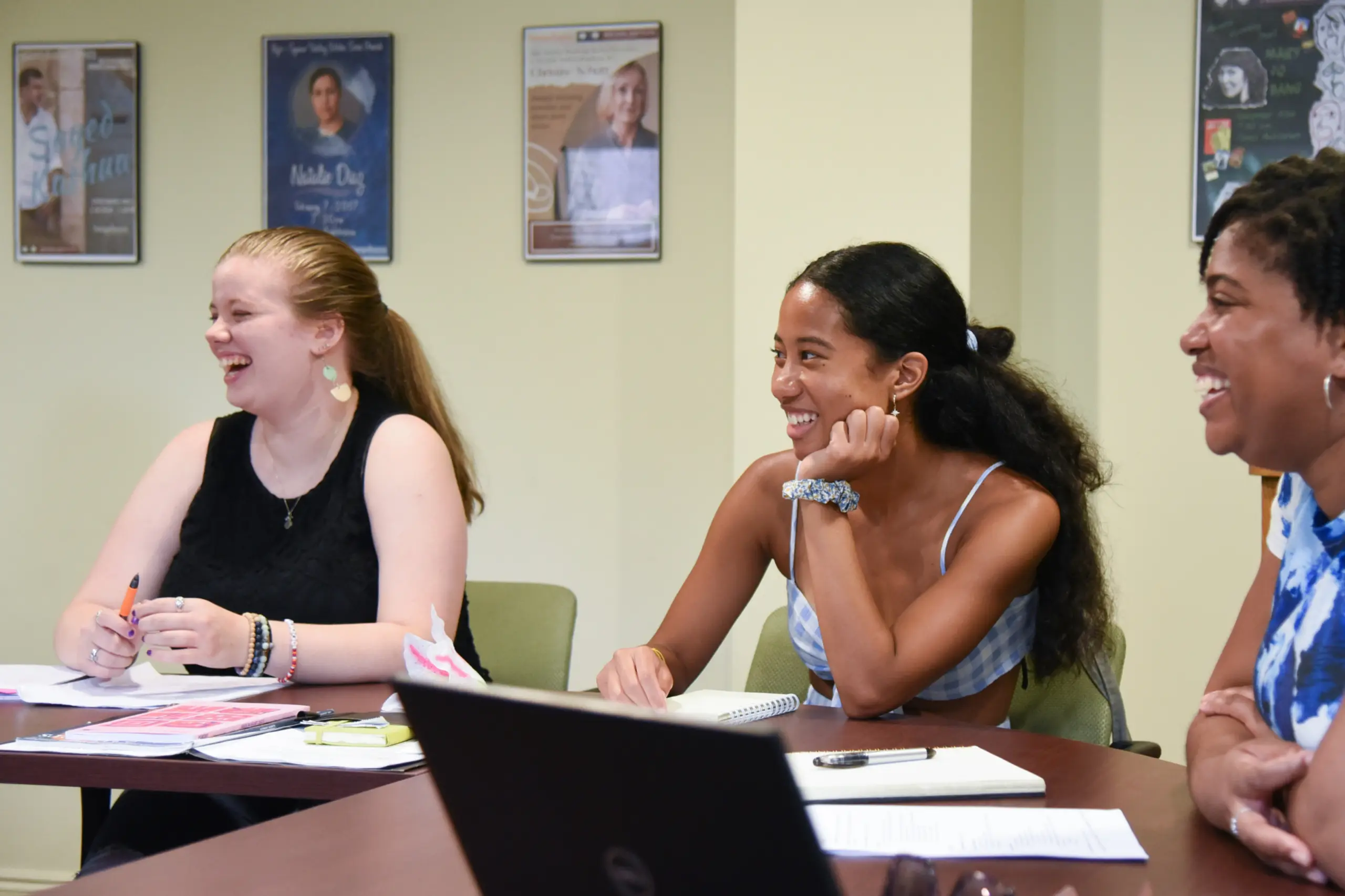 Three women sit at a table laughing during a meeting or class, with notebooks, papers, and pens in front of them. Posters are visible on the wall in the background.