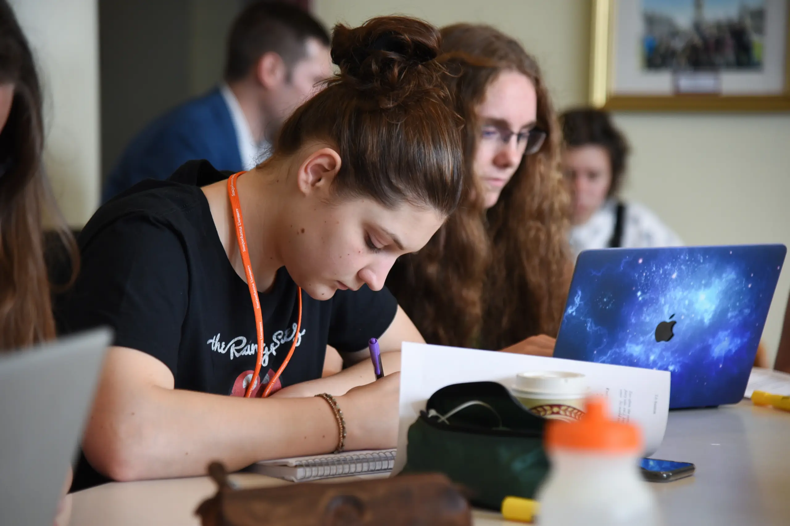 A young woman with brown hair tied up writes in a notebook at a table, surrounded by papers. Others around her are focused on their own work, with one person using a laptop featuring a space-themed cover.