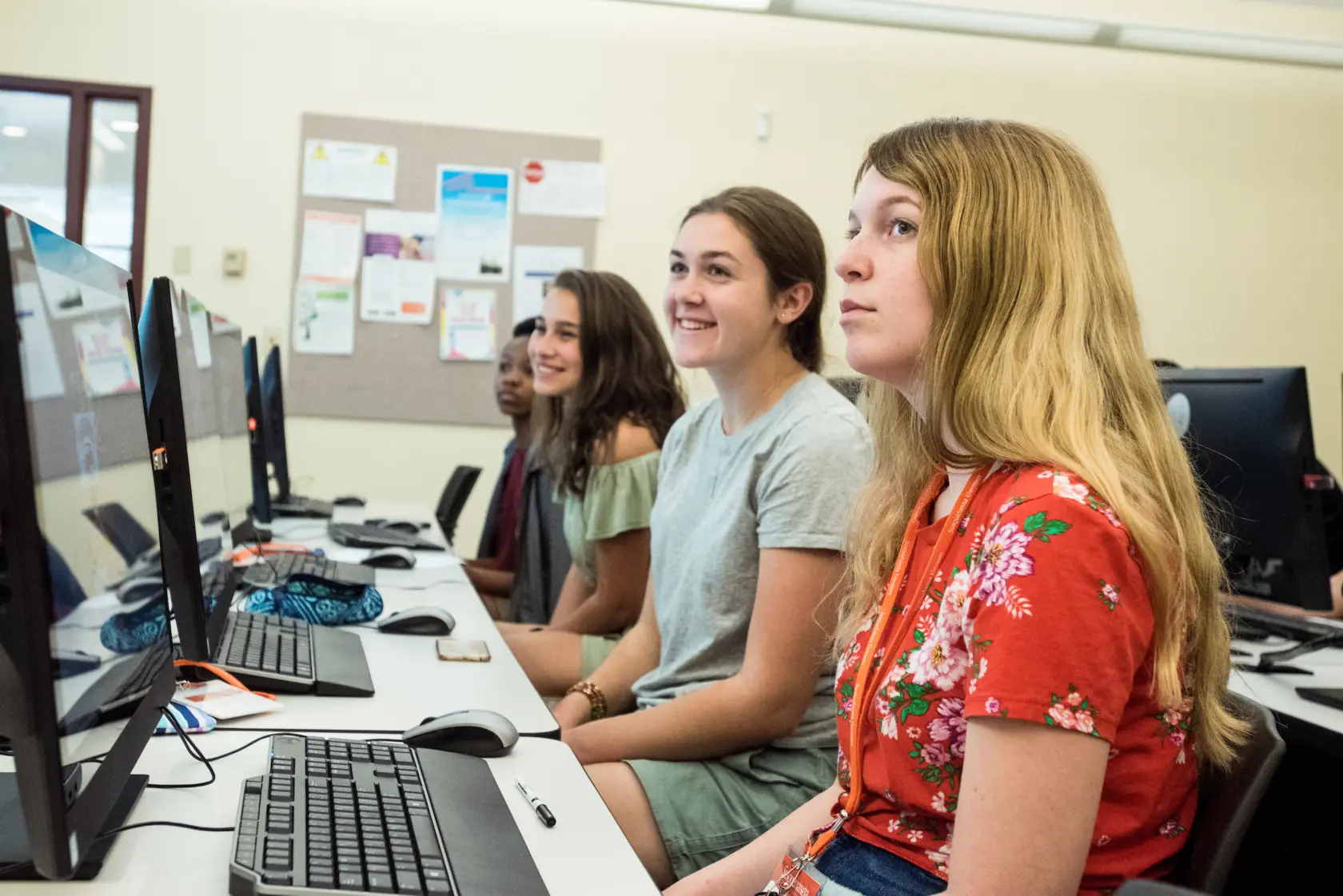 Four young women sit in front of desktop computers in a classroom, attentively looking at their screens. The classroom has a bulletin board with papers on the wall in the background.
