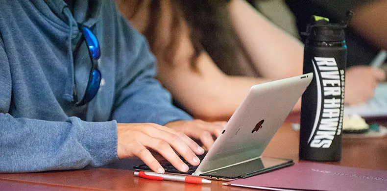 A person wearing a blue hoodie types on a silver laptop at a desk. A black water bottle labeled "River Hawks" and a red pen are visible nearby. Others are sitting and writing in the background.