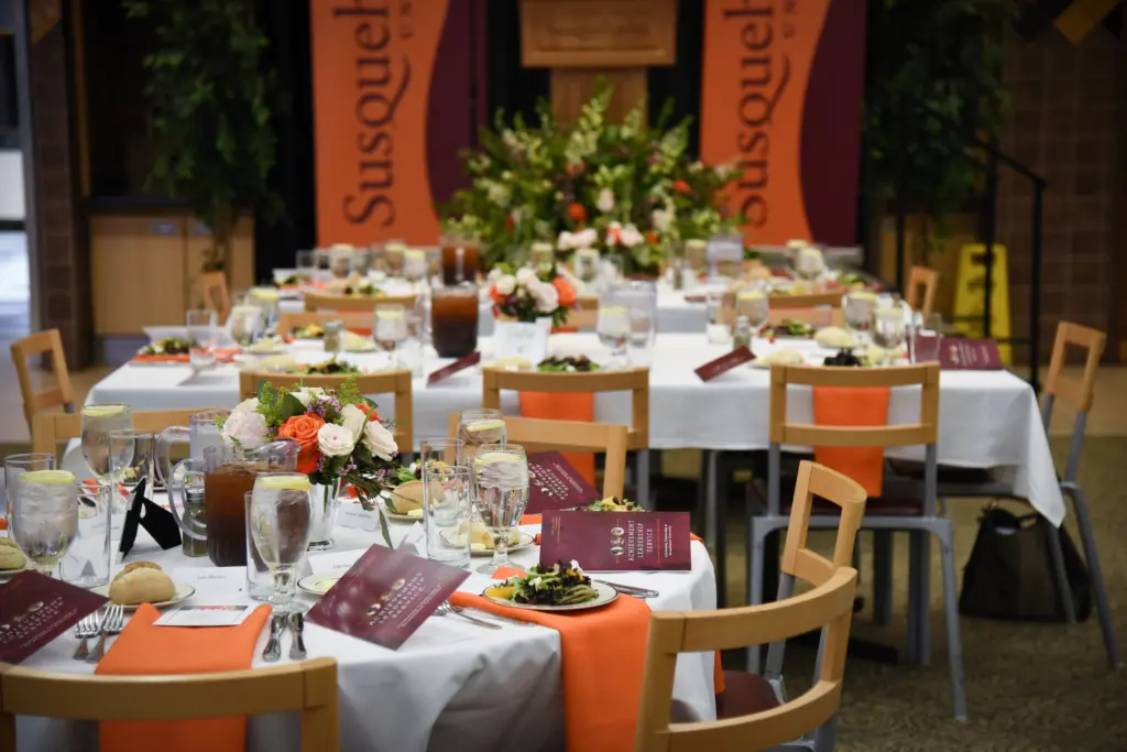 A banquet setup with tables covered in white cloths, orange napkins, flower centerpieces, salads, drinks, and menus. In the background, banners reading “Susquehanna,” a large floral arrangement, and signs for Conference Facility Rental are visible.
