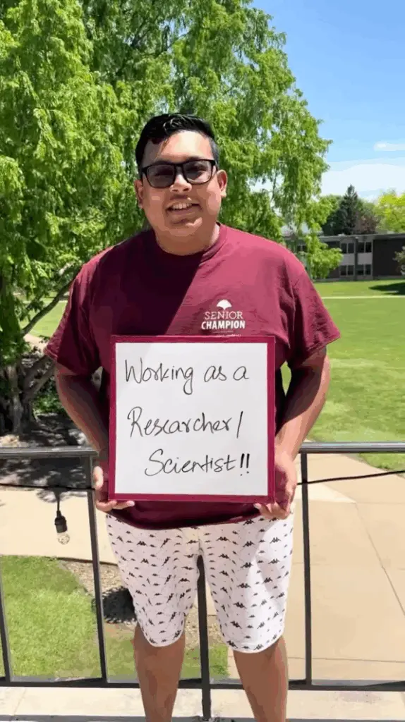 A smiling person stands outdoors holding a sign that says, "Working as a Researcher! Scientist!!" They wear glasses, a maroon shirt, and white shorts with patterns. Green trees and grass are visible in the background.